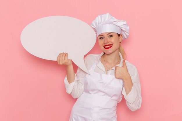 Vista frontal confitería femenina en ropa blanca con cartel blanco y sonriendo en la pared rosa trabajo de pastelería dulce de confitería