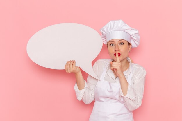 Vista frontal confitería femenina en ropa blanca posando con letrero blanco que muestra el signo de silencio en la pared rosa confitería trabajo de pastelería dulce