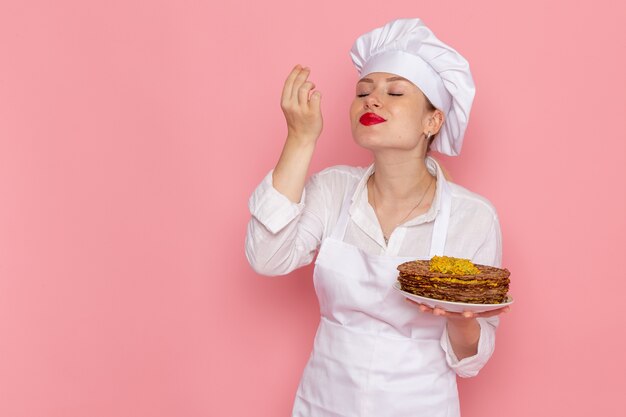 Vista frontal confitería femenina en ropa blanca sosteniendo deliciosos pasteles en la pared rosa trabajo de pastelería dulce confitería trabajo