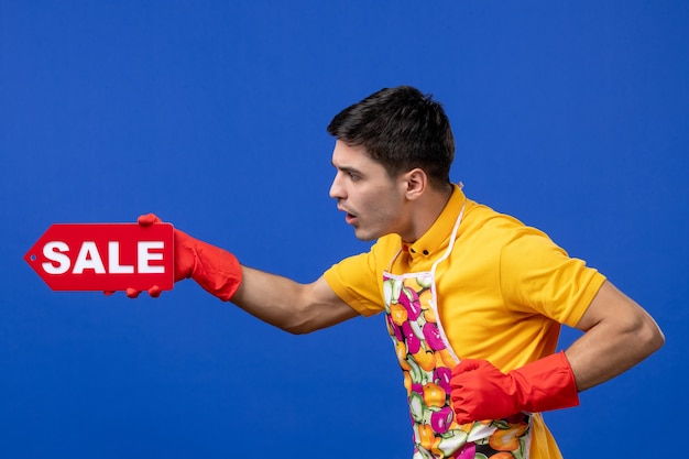 Vista frontal confundida ama de llaves masculina en camiseta amarilla con cartel de venta en espacio aislado azul