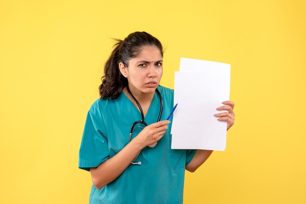 Vista frontal confundida bastante doctora sosteniendo papeles y bolígrafo sobre fondo amarillo