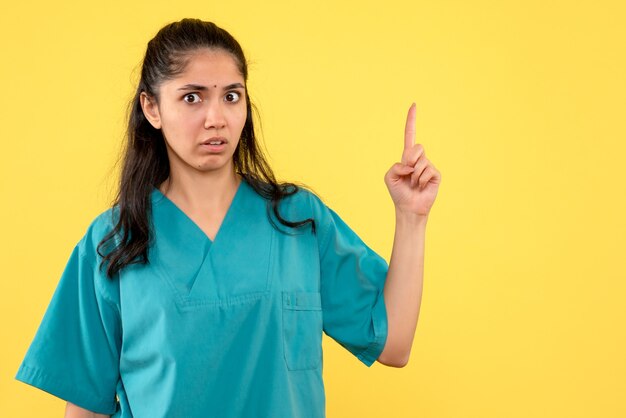 Vista frontal confundida doctora en uniforme apuntando con el dedo hacia arriba sobre fondo amarillo