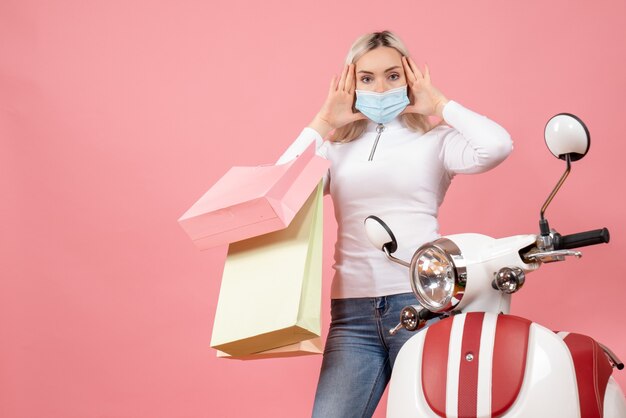 Vista frontal confundida jovencita sosteniendo bolsas de compras cerca de ciclomotor