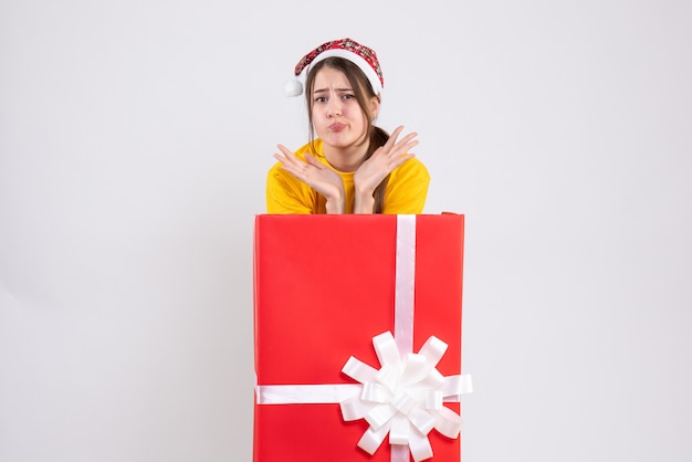 Vista frontal confundida linda chica con sombrero de santa de pie detrás de un gran regalo de Navidad