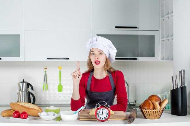 Vista frontal confundida mujer rubia con sombrero de cocinero y delantal apuntando al techo en la cocina