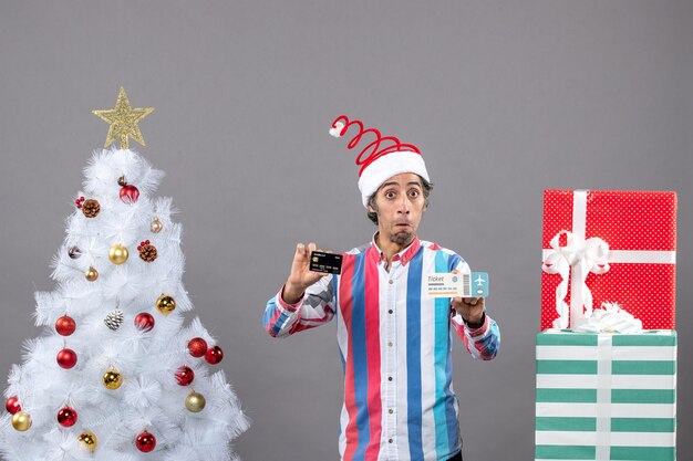 Vista frontal confundido hombre que sostiene la tarjeta y el boleto de viaje alrededor del árbol de Navidad y regalos