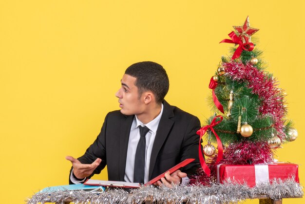 Vista frontal confundido hombre en traje sentado en la mesa sosteniendo el documento mirando a la derecha árbol de Navidad y regalos