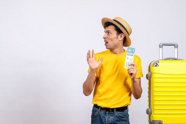 Vista frontal confundido joven en camiseta amarilla de pie cerca de la maleta amarilla con boleto
