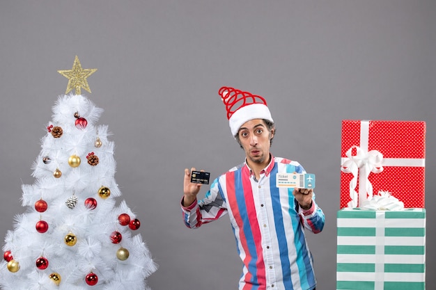 Vista frontal confundido joven sosteniendo la tarjeta y el boleto de viaje alrededor del árbol de Navidad y regalos