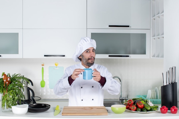 Vista frontal confuso chef masculino con sombrero de cocinero sosteniendo la taza azul de pie detrás de la mesa de la cocina