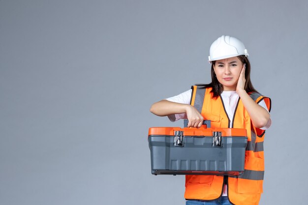 Vista frontal del constructor femenino con caja de herramientas pesada en pared gris