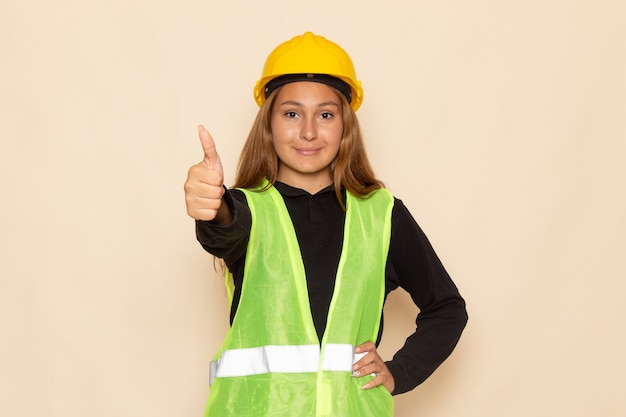 Vista frontal del constructor femenino en casco amarillo camisa negra sonriendo mostrando como signo en la pared blanca