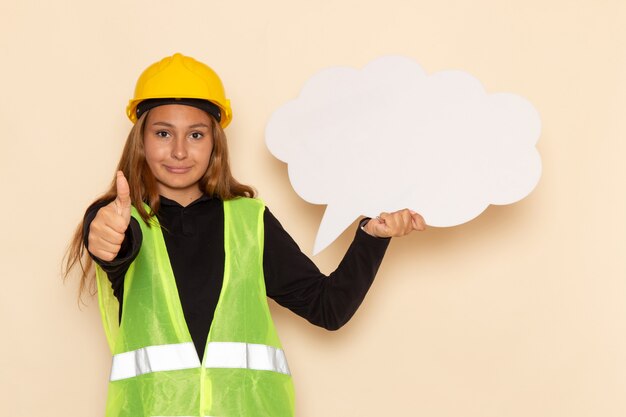 Vista frontal del constructor femenino en casco amarillo con cartel blanco y sonriendo en la pared blanca construcción de constructor femenino