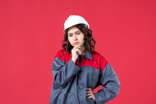 Vista frontal del constructor femenino pensativo en uniforme con casco sobre fondo rojo aislado