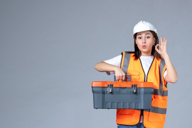 Vista frontal del constructor femenino que lleva la caja de herramientas pesadas en la pared blanca