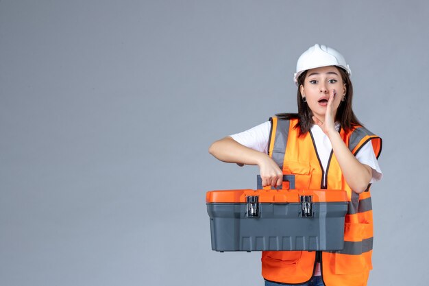 Vista frontal del constructor femenino que lleva la caja de herramientas pesadas en la pared blanca