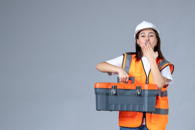 Vista frontal del constructor femenino que lleva la caja de herramientas pesadas en la pared blanca