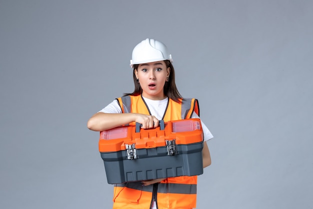 Vista frontal del constructor femenino sosteniendo la caja de herramientas en la pared gris