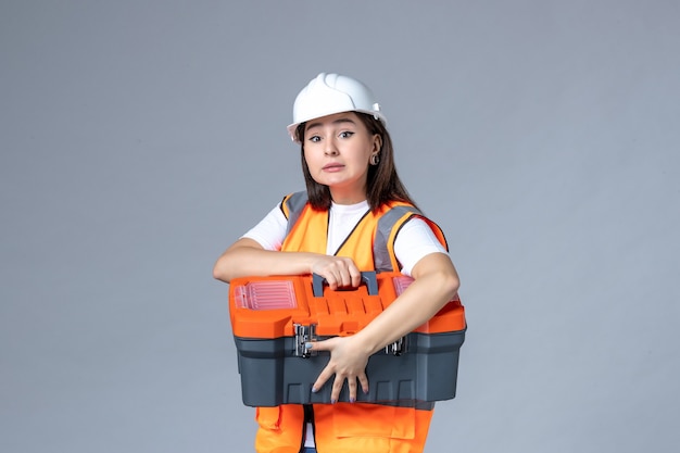 Foto gratuita vista frontal del constructor femenino sosteniendo la caja de herramientas en la pared gris