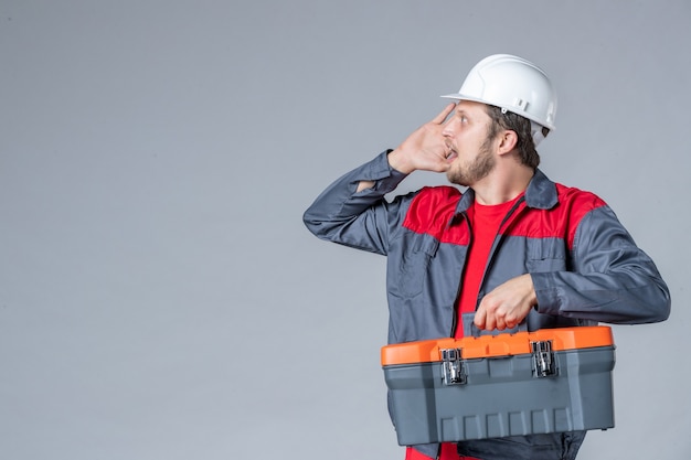 Vista frontal del constructor masculino en caja de herramientas de sujeción uniforme sobre fondo gris