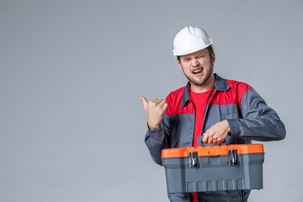 Vista frontal del constructor masculino en caja de herramientas de sujeción uniforme sobre fondo gris