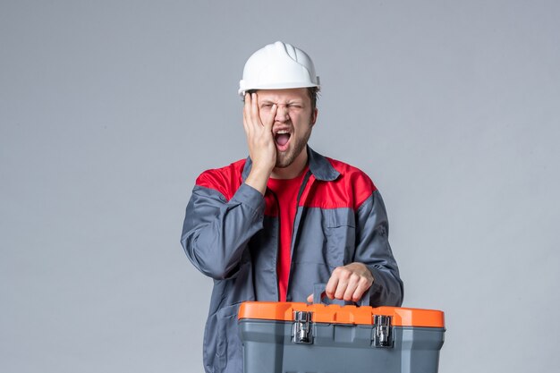 Vista frontal del constructor masculino en caja de herramientas de sujeción uniforme sobre fondo gris