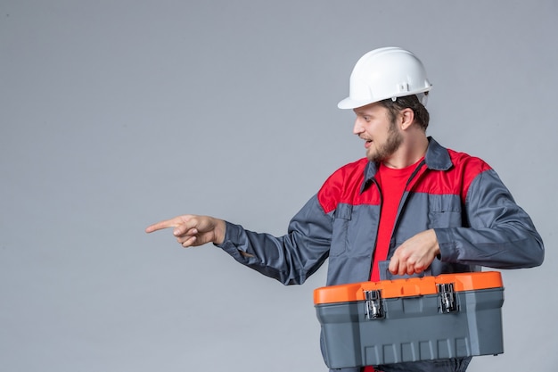 Vista frontal del constructor masculino en caja de herramientas de sujeción uniforme sobre fondo gris