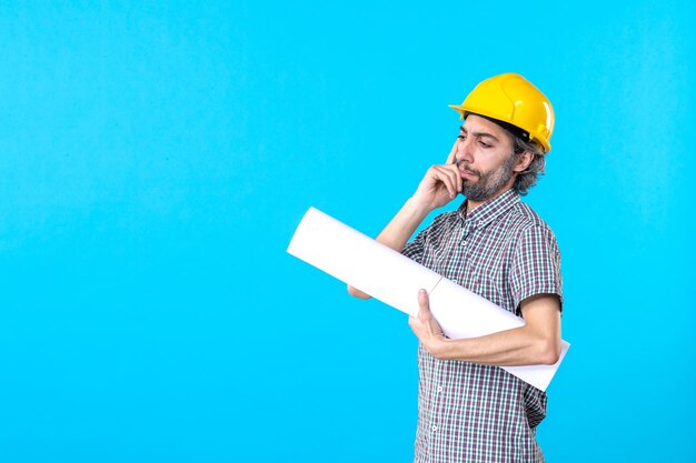 Vista frontal del constructor masculino en casco amarillo con plan sobre un fondo azul, constructor de arquitectura de propiedad, trabajador, ingeniero de trabajo de construcción