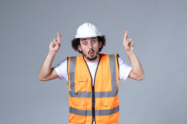 Vista frontal del constructor masculino emocional conmocionado con chaleco de advertencia con casco de seguridad y cruzando los dedos sobre la pared gris de la ola