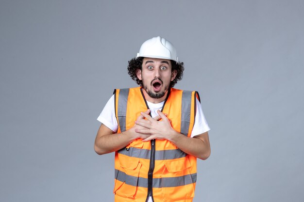 Vista frontal del constructor masculino emocional conmocionado vistiendo chaleco de advertencia con casco de seguridad en la pared de onda gris