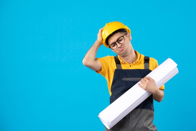 Vista frontal del constructor masculino estresado en uniforme amarillo con plan de papel en azul