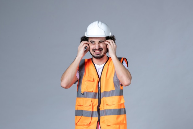 Vista frontal del constructor masculino incierto inseguro con chaleco de advertencia con casco de seguridad en la pared de onda gris