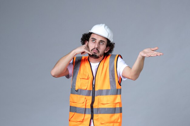 Vista frontal del constructor masculino inseguro e incierto con chaleco de advertencia con casco de seguridad y haciendo el gesto de llamarme en la pared de onda gris