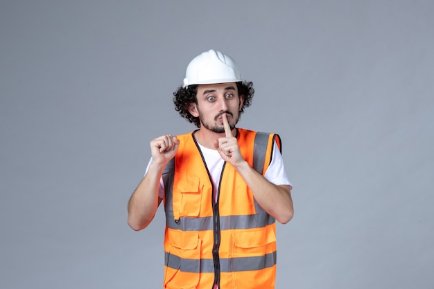 Vista frontal del constructor masculino nervioso vistiendo chaleco de advertencia con casco de seguridad y haciendo gesto de silencio en la pared de onda gris