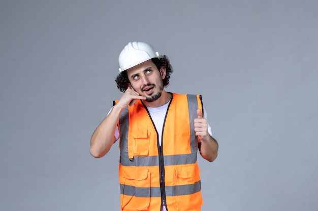 Vista frontal del constructor masculino ocupado vistiendo chaleco de advertencia con casco de seguridad y haciendo gesto de llamarme en la pared de onda gris