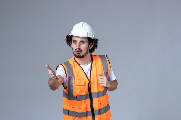 Vista frontal del constructor masculino preguntándose con chaleco de advertencia con casco de seguridad en la pared de onda gris