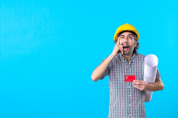 Vista frontal del constructor masculino que sostiene el plan y la tarjeta bancaria sobre fondo azul, diseño de dinero, trabajo de constructor, trabajo, edificios de arquitectura de color