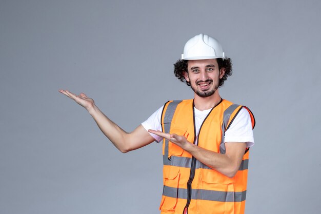 Vista frontal del constructor masculino sonriente con chaleco de advertencia con casco de seguridad y apuntando algo en el lado derecho en la pared de onda gris