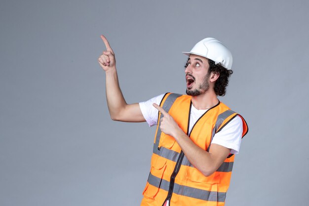 Vista frontal del constructor masculino sonriente vistiendo chaleco de advertencia con casco de seguridad y apuntando hacia arriba en la pared de onda gris