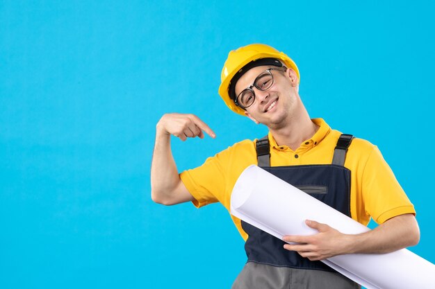 Vista frontal del constructor masculino en uniforme amarillo y casco con plan en pared azul