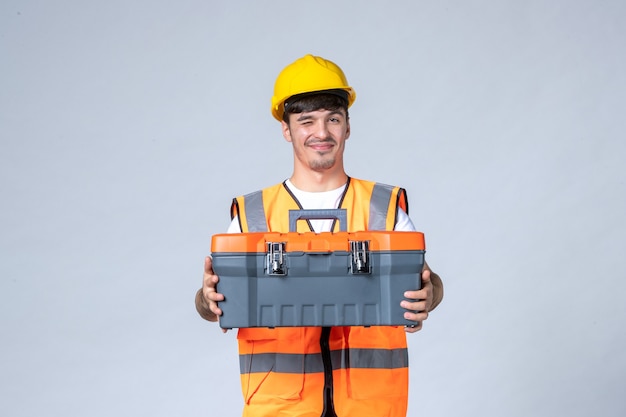 Vista frontal del constructor masculino en uniforme y casco amarillo con caja de herramientas en la pared blanca