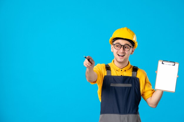 Vista frontal del constructor masculino en uniforme y casco con bloc de notas en la pared azul