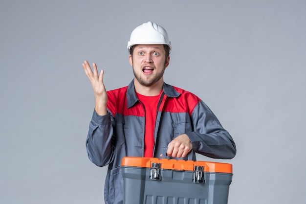 Vista frontal del constructor masculino en uniforme y casco con maletín de herramientas sobre fondo gris