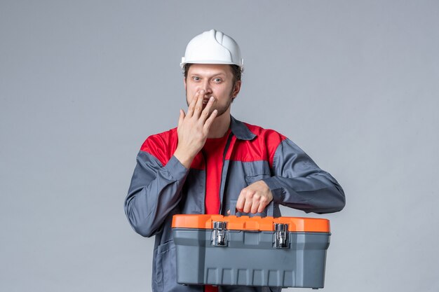 Vista frontal del constructor masculino en uniforme y casco con maletín de herramientas sobre fondo gris