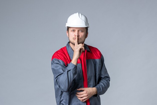 Vista frontal del constructor masculino en uniforme y casco pidiendo silencio sobre fondo gris