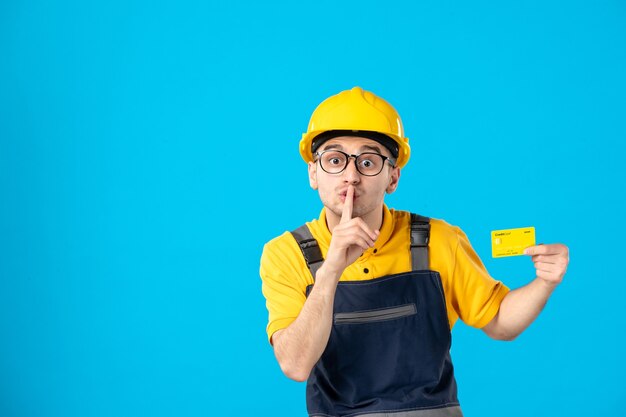 Vista frontal del constructor masculino en uniforme y casco con tarjeta de crédito pidiendo silencio en la pared azul