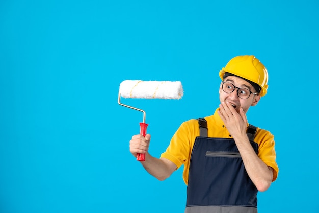 Vista frontal del constructor masculino en uniforme con rodillo de pintura en sus manos sobre la pared azul