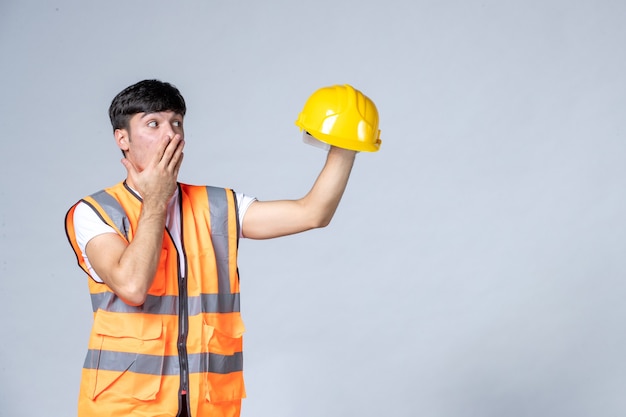 Vista frontal del constructor masculino en uniforme sosteniendo casco amarillo en la pared blanca