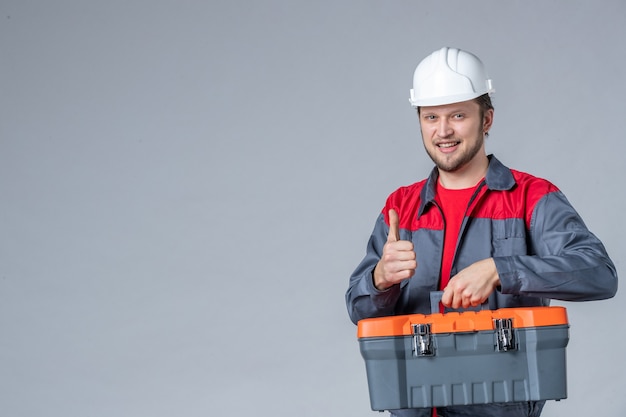 Foto gratuita vista frontal del constructor masculino en uniforme sujetando la caja de herramientas con una sonrisa sobre fondo gris