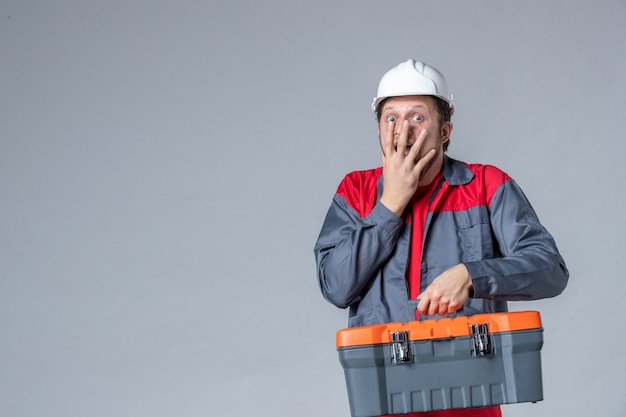 Vista frontal del constructor masculino en uniforme sujetando la caja de herramientas sorprendido y emocionado sobre fondo gris